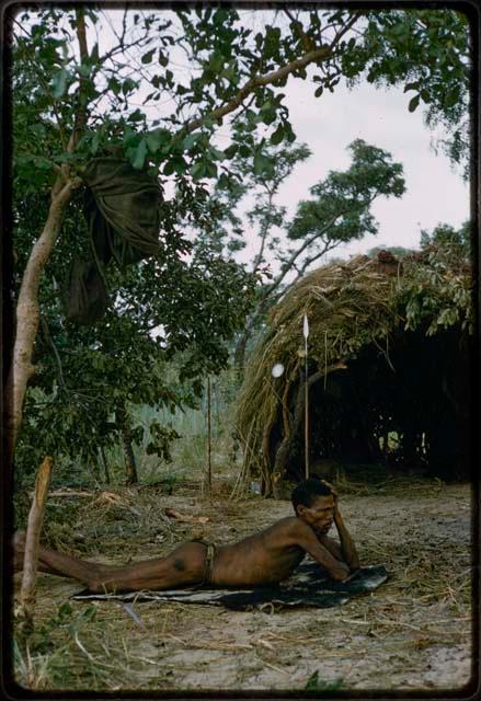 "Gao Medicine" lying on his stomach in front of a skerm, with his belongings hanging on a tree