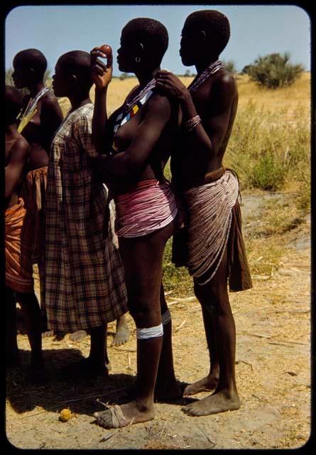 Group of women and girls standing, including two women wearing bead aprons dyed pink, with a girl wearing a dress standing next to them