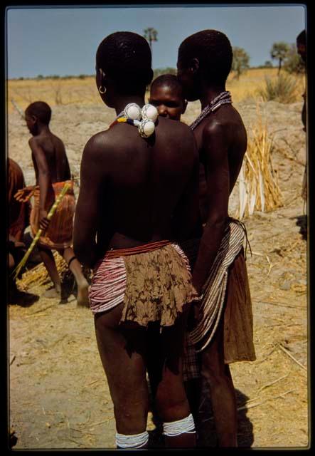 Two women wearing bead aprons, one wearing a necklace of big shells, view from behind