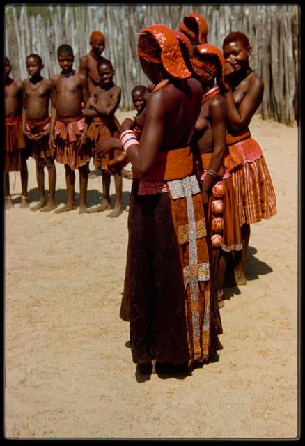 First wife of Chief Nehemiah wearing an ornamented skirt and a pink headdress, view from behind