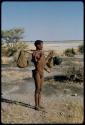 Carrying: /Ti!kay carrying bags of nuts and Ovambo pot on a carrying stick, using his assegai over the left shoulder to distribute the weight, from the side, with Gautscha Pan in the background