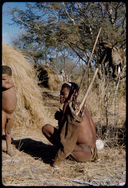 Carrying: Man holding a bag containing a bow and long sticks on his shoulder, sitting next to a skerm, with a child standing next to him