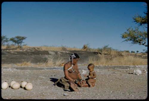 Children, Eating: !U sitting by the pan holding !Ungka Norna (after her hair was cut) on her lap, with ostrich egg shells beside her