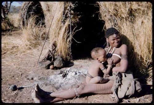 Children, Mothers nursing babies: Woman with scarification leaning against a skerm, nursing a child; showing belongings in bags, baby's haircut and sinew wrapped around body