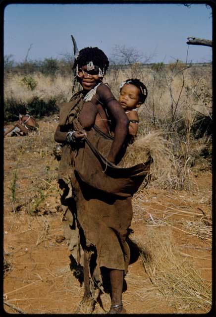 Children, Carrying: !Ghia standing, with her baby tied to her back and carrying grass in her kaross