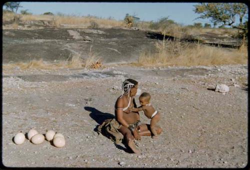 Children, Norna: !U sitting with !Ungka Norna standing against her near a waterhole, with ostrich egg shells on the ground next to them