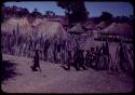 Women walking, carrying baskets on their heads, with huts in the background