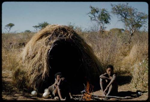 Marriage: N!ai sitting with /Gunda next to a fire in front of their skerm, after she went out to gather veldkos