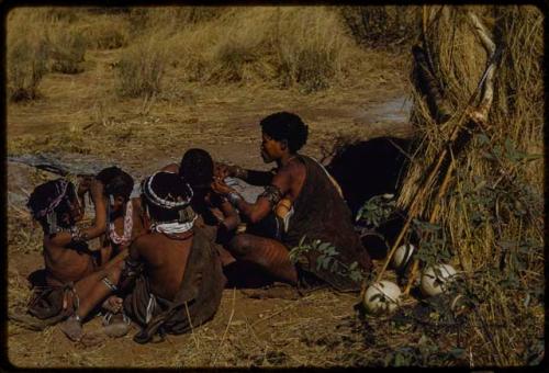 Ornament: Group of women sitting closely together in front of a skerm, with Khuan//a fixing another woman's earring