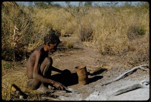 Portraits: "Old /Gishay" sitting next to the ashes of a fire, with a wooden container on the ground next to him; he is scraping the ashes with something