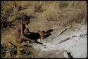 Portraits: "Old /Gishay" sitting next to the ashes of a fire, with a wooden container on the ground next to him; he is scraping the ashes with something