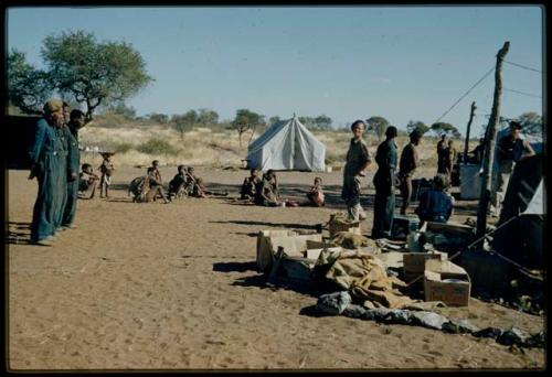 Expedition, Gifts: Group of women sitting in the expedition camp, with Franz Rudolph standing on the left, John Marshall standing in the middle, Kernel Ledimo standing next to Lorna Marshall sitting, and Heinrich Kretzchmar standing on the right