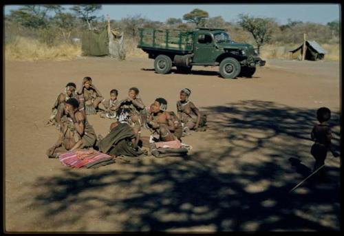 Expedition, Gifts: Group of women sitting in the expedition camp, with cloth given to them by expedition members on the ground next to them, expedition truck and sound equipment tent in the background