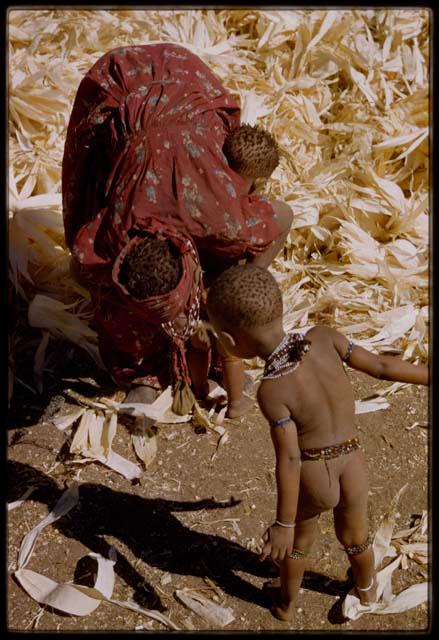 Expedition, Metzger: Woman bending over to wipe her baby, another child watching, with a pile of corn husks in the background