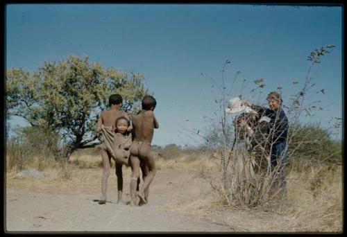 Expedition, Photography: Two boys carrying "Little ≠Gao" in a kaross, playing the "airplane game," with Lorna Marshall standing near them holding her helmet over a camera lens