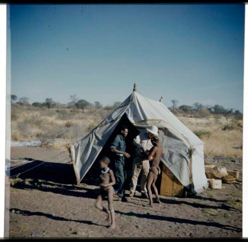 Expedition, Ethnographic work: Person receiving something from Laurence Marshall in front of the expedition storage tent, with another expedition member standing next to them and a child walking away