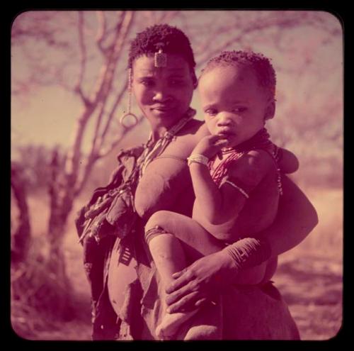 Children, Mother and children: Woman wearing fiber bracelets, copper ring on a string of beads in her hair, a triangular ostrich eggshell bead ornament, and sha sha (root used in the choa ceremony), standing and holding a baby