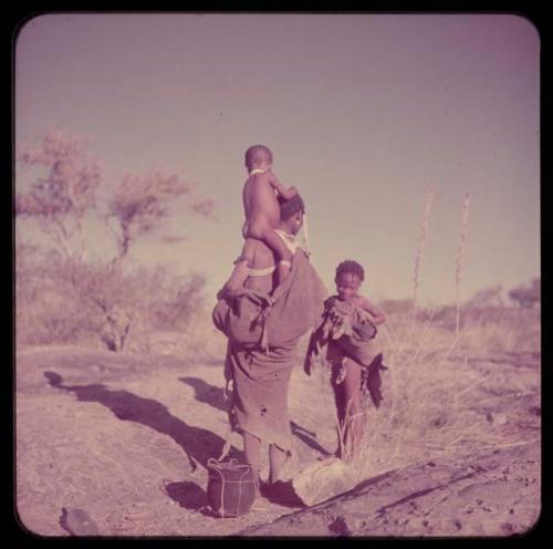 Children, Carrying: /Naoka standing at a waterhole, carrying a child on her shoulders, with a girl standing nearby and an Ovambo pot on the ground