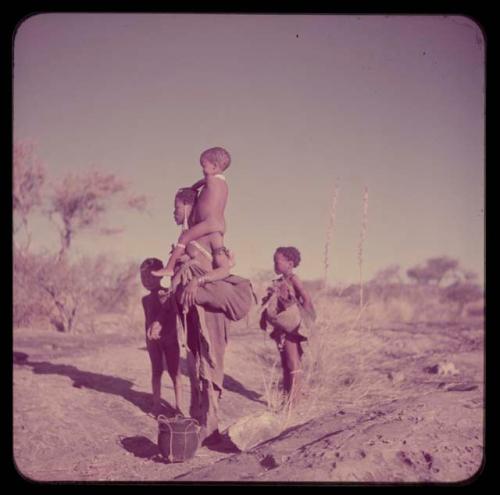 Children, Carrying: /Naoka standing at a waterhole, carrying a child on her shoulders, with !Nani and an unidentified girl standing nearby and an Ovambo pot on the ground