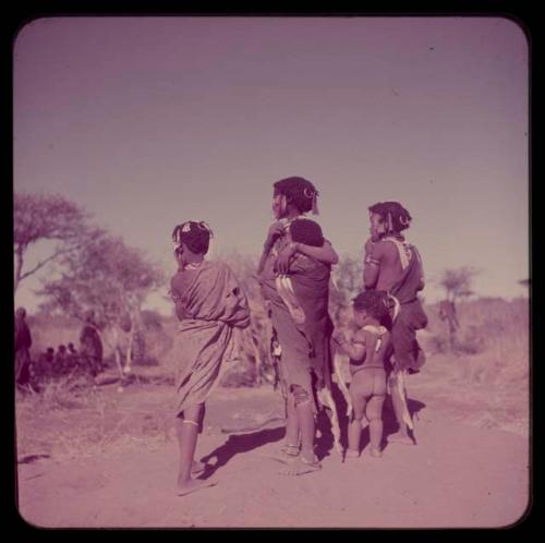 Children, Carrying: Girls standing, one wearing beads and carrying a baby, view from behind