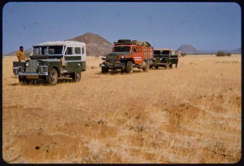 Expedition trucks parked in an area with scant grass, hills in the background