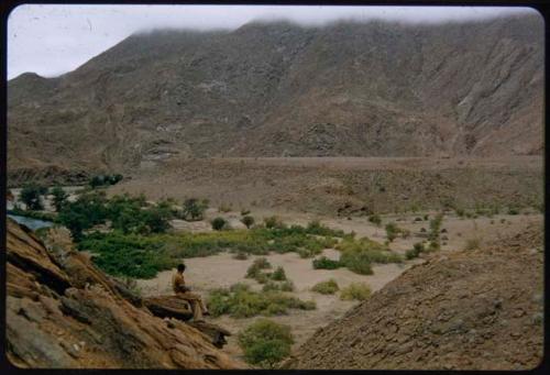 Heinrich Neumann sitting on a rock overlooking trees next to the Kunene River, with huts in the background