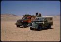 Expedition trucks (Land Rover and Dodge) on a flat plain, with sand dunes in the background