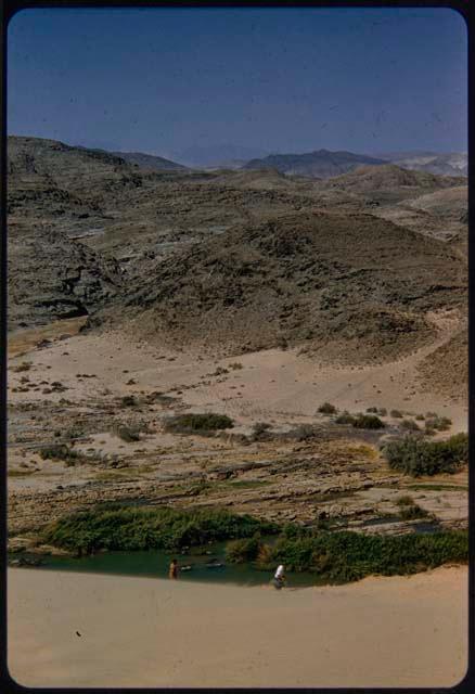 People walking along the edge of a sand dune next to the Kunene River, with hills in the background