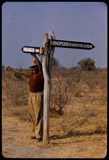 Philip Hameva standing under a signpost and pointing toward "Ondangua" (Ondangwa)