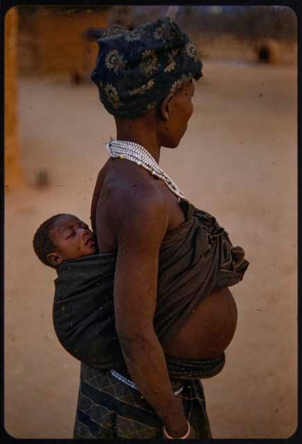 Woman standing, with a baby tied to her back, profile