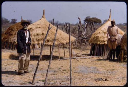 Shikongo, headman at Mulungo, and another man standing next to huts and storage baskets