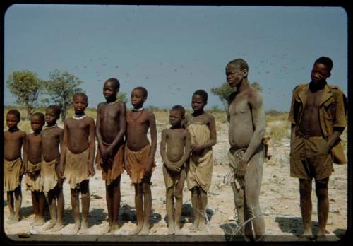 Group of children standing in a line, with two men standing next to them, one covered in mud