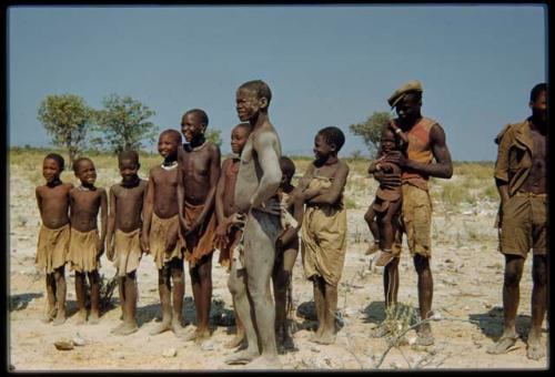 Group of children standing in a line, with three men standing next to them, one covered in mud