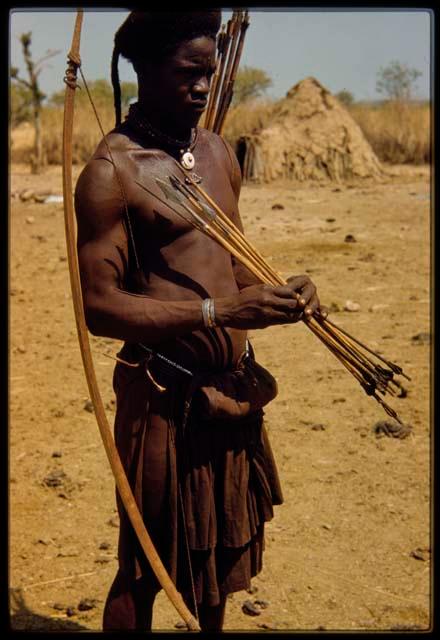 Hunter carrying a bow over his shoulder, holding arrows with metal tips and others with poison applied