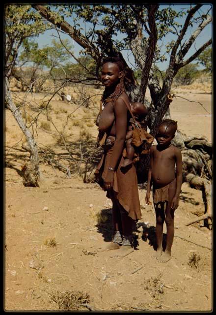 Woman with baby tied to her back, standing with a child in front of a tree