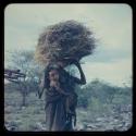 Woman carrying bundles of grass over her head, with a baby tied to her side
