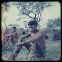 "Gao Short" sitting in the grass after a curing dance, with other people sitting in the background