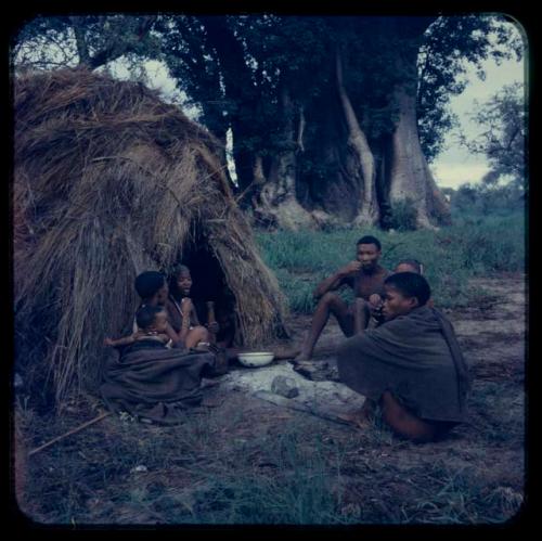 Woman sitting in a skerm and pounding something in a mortar, with other people sitting opposite her