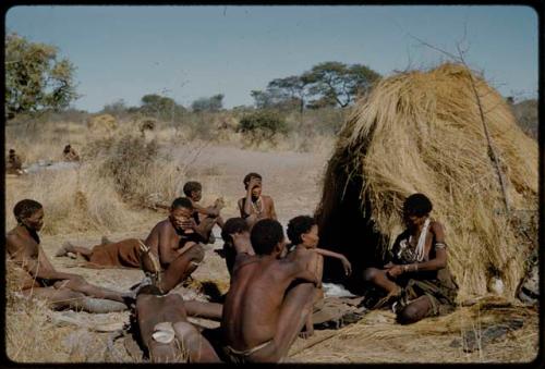 Sinue [sinew]: Khwo//o-/Gasa making a cord, sitting in front of a skerm with a group of people including ≠Gao (her husband) with his hand over his face and /Naoka ("Gao Medicine's" wife) with her arm on her knee