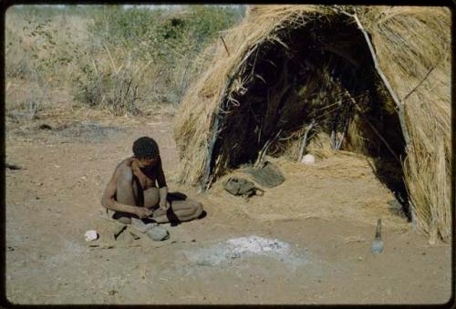 Skerm: Man working on something in front of a skerm, holding his stone anvil with his foot, with a grass bed and an ostrich egg inside the skerm