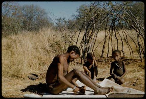 Scraping Skin: /Ti!kay scraping a skin, with two boys sitting next to him