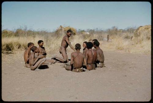 Story Telling, Game: Group of men and boys playing !’Om g!xuni / ’Ai kota !U!u g!xuni / N!aih //’an (the porcupine game, or "axe and assegai game") with "Gao Medicine" on his knees and /Gunda smiling up at him