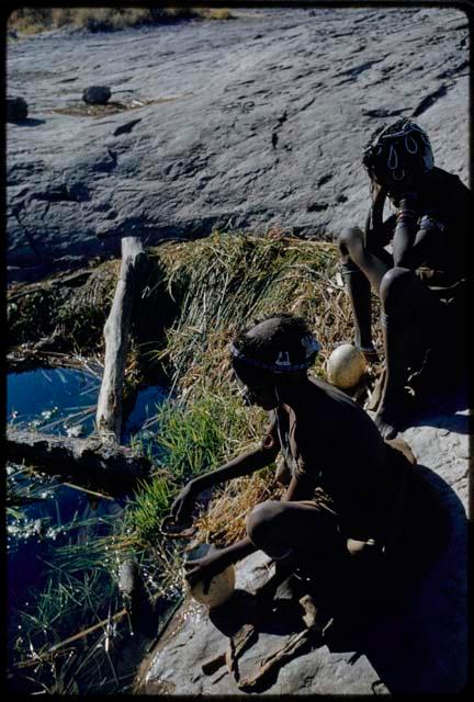 Water: Bau (daughter of Ti!kay) filling an ostrich eggshell with a tortoise shell dipper, with another woman sitting next to her, view from the side