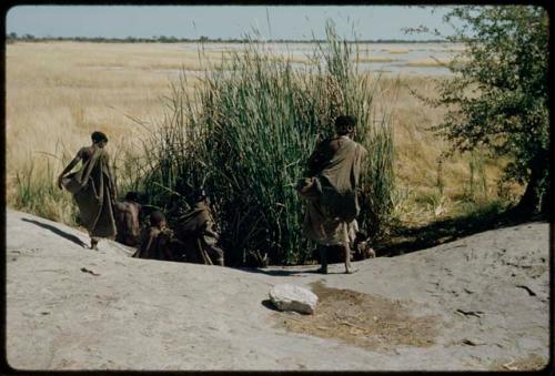 Water: People standing and sitting around a waterhole, seen from a ledge, with a pan filled with water in the distance