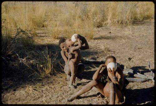 Water: /Gunda (N!ai's husband) helping a child to drink from an ostrich eggshell, with another person sitting and drinking next to them