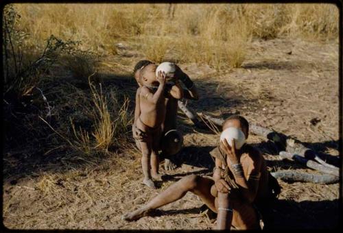 Water: /Gunda (N!ai's husband) helping a child to drink from an ostrich eggshell, with another person sitting and drinking next to them