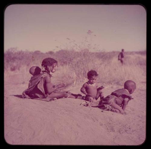 Children, Music: N!ai sitting on a termite mound playing a //guashi, with !Ungka Norna tied to her back, Bau and /Gaishay (Di!ai's son) sitting next to her