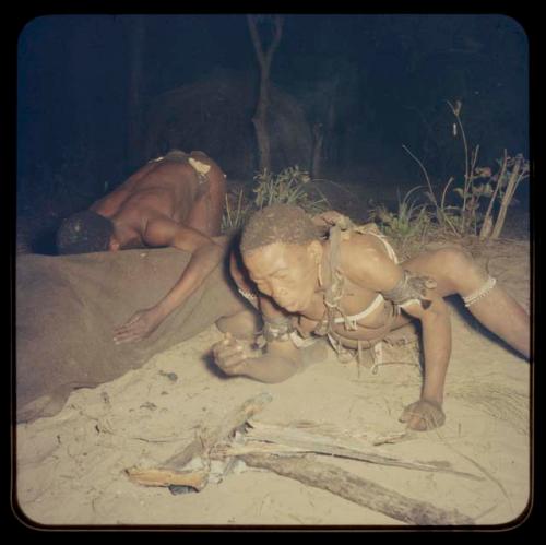 Medicine man blowing on fire, with another man leaning over in the background