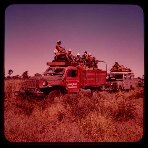 Group of people riding in trucks on the way from !O to !Nama; the red truck reads "Peabody Museum of Harvard University" on its front door