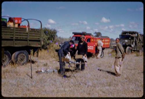 Wilhelm Camm, Dabe, and a mechanic standing by chairs, expedition trucks in the background